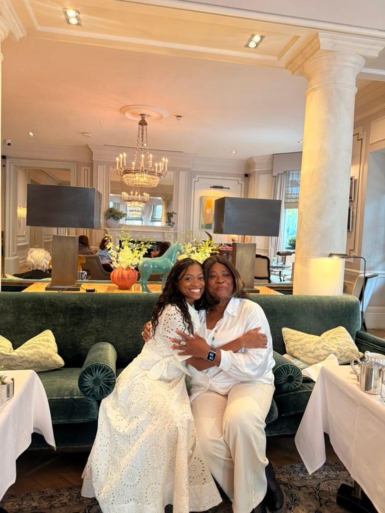 Nadia and her mum sit together on a green velvet sofa, smiling and embracing in an elegant lounge decorated with chandeliers, flowers, and artwork.