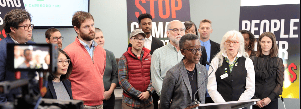 A wide view of members of Carrboro Town Council and others standing behind Mayor Barbara Foushee at a press conference. Banners behind the people are mostly obscured, with the words