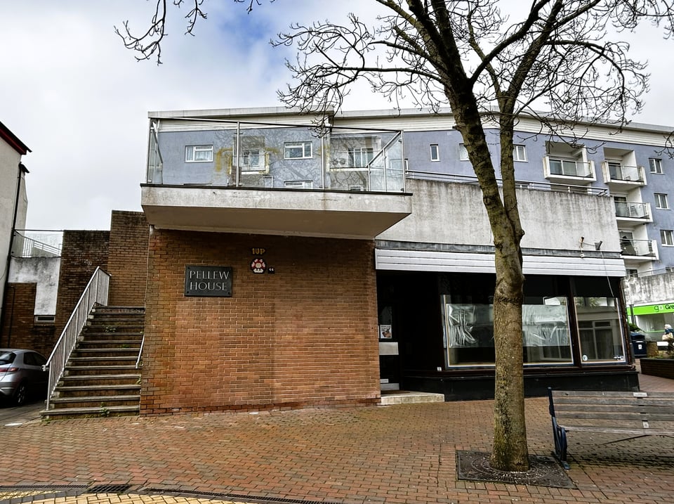 A five storey block of flats with pale bluegrey render. In front of them and at right angles is a large brick single storey building with concrete stairs up to an open roof. A slate sign reads 'Pellew House' but the white render parapet is discoloured and the ground floor shop is empty.