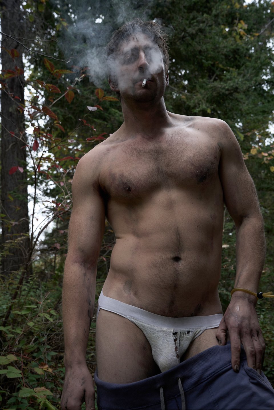 man in a torn jockstrap smokes a cigarette as he stands in the trees