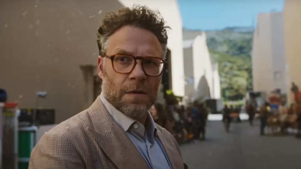 Seth Rogen, a white man with short curly hair and a graying beard, wearing red-rimmed glasses and a tweed jacket, stands on a blurry studio lot.