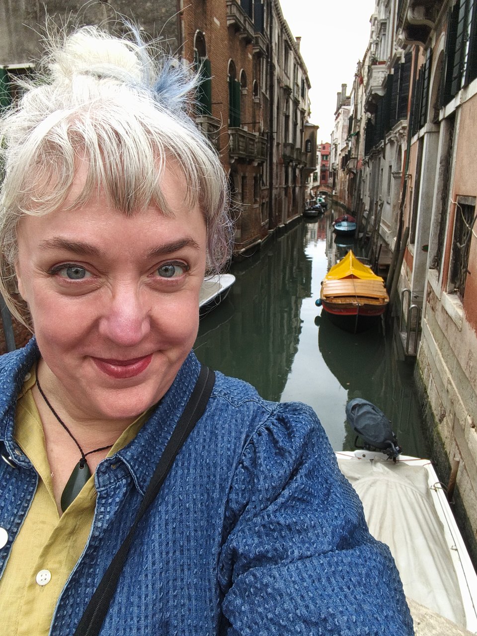 an image of Rachael, a white/blue haired white woman grinning rather maniacally above a canal on a cloudy day