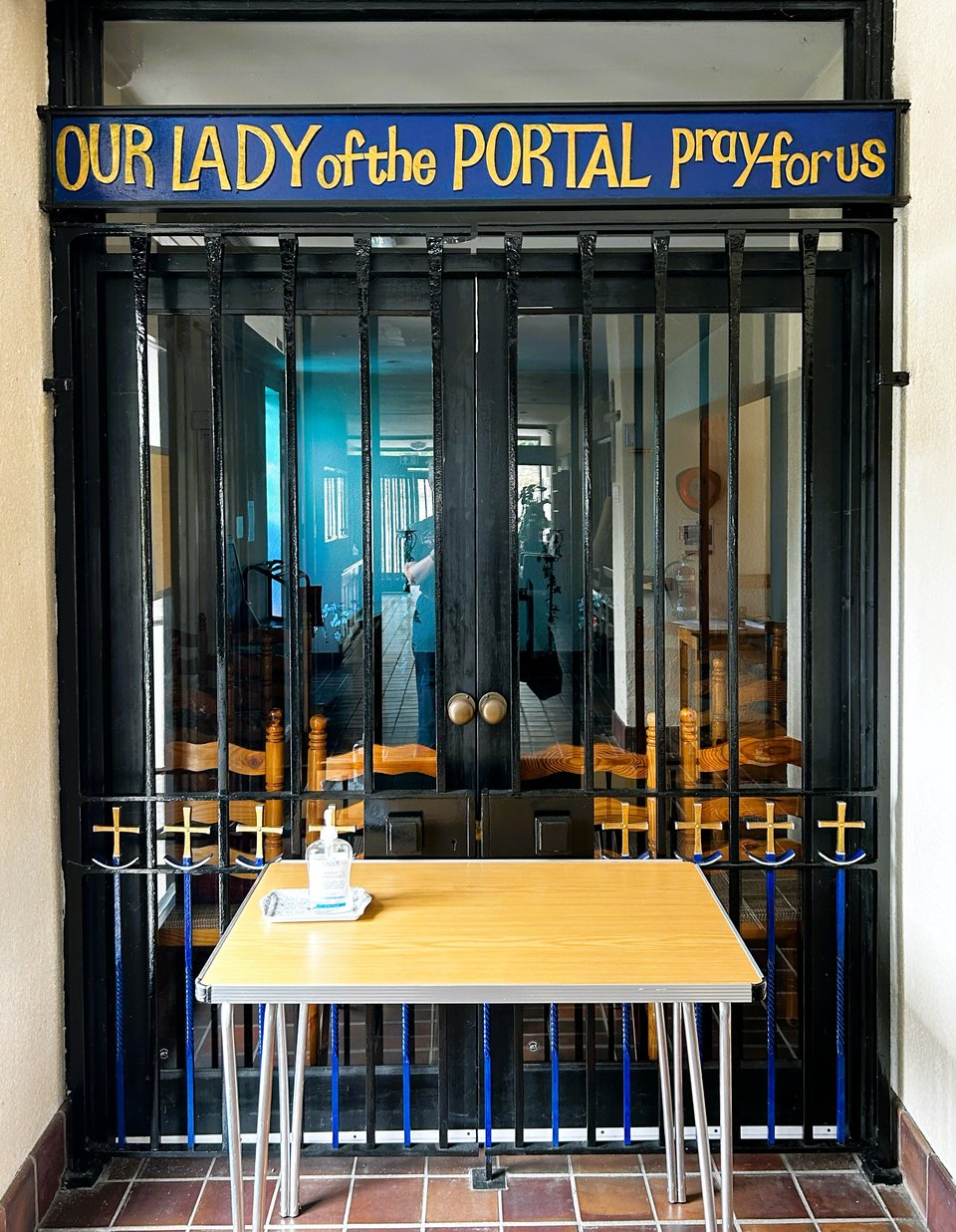 Black gates with a simple blue and gold detail at the bottom which is mostly hidden by a table holding some hand sanitiser. Above the gate, in lovely 1970s gold font on a blue background are the words Our Lady of the Portal pray for us.