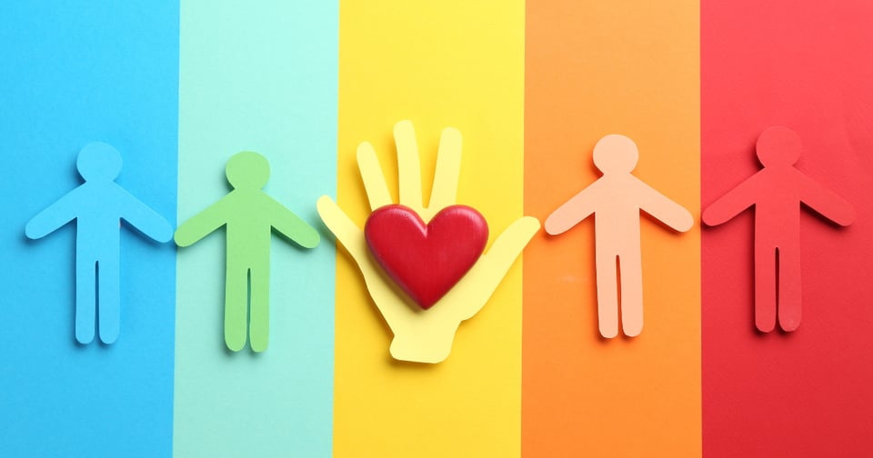 Image of a rainbow background. On top are paper cutouts of people. At the center is a yellow hand with a red heart on it.
