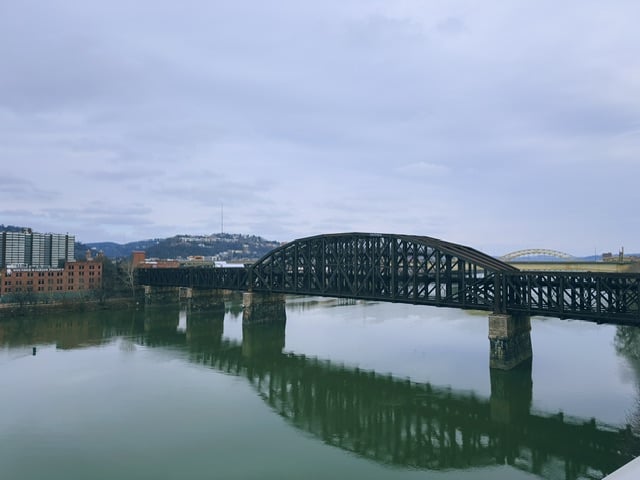 An old, steel train bridge crossing one of the Pittsburgh rivers. In the background is a newer, yellow steel bridge for vehicles. The train bridge is reflected in the river.