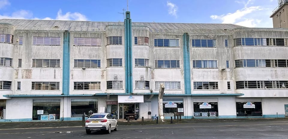 A four storey art deco building. The ground floor is plate glass shop windows. The upper storeys have lots of critall windows, including ribbon ones around the curving bays. A short fin sticks up in the centre. The white render and sky blue details are very very grubby.