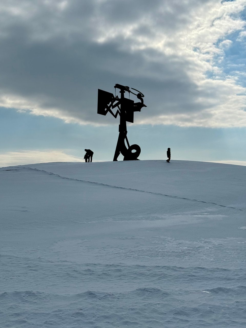 photo of an abstract sculpture atop a snow-covered hill, with a man and two boys playing beside it in the snow