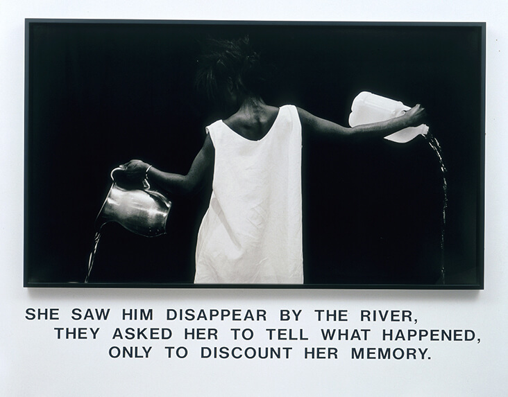 image description: Lorna Simpson, Waterbearer. features a black woman in a simple white gown, seen from the back, pouring water from a metal jug in one hand and a plastic bottle in the other.