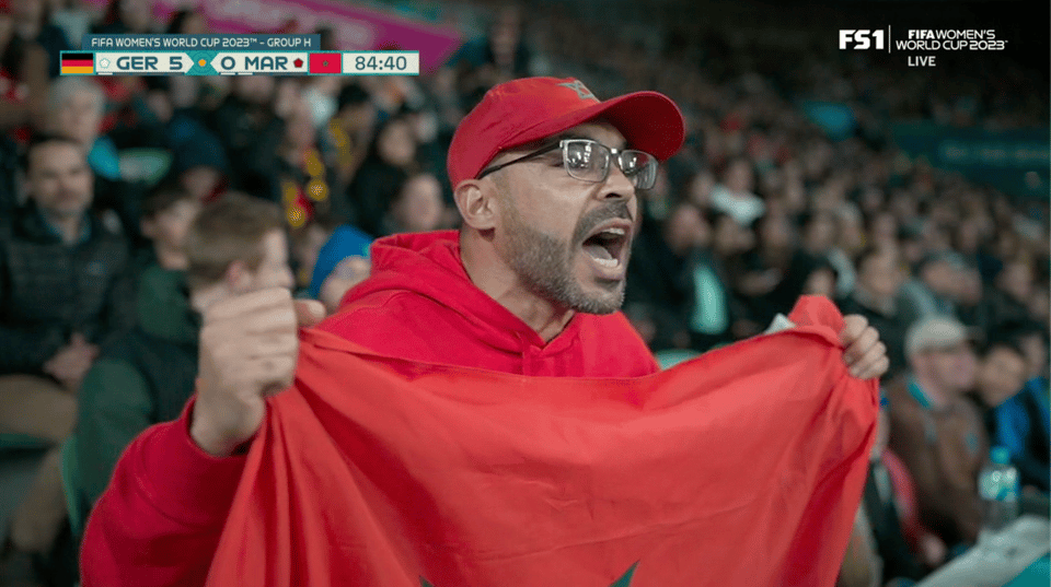 A Morocco fan clad in all red, holding the country's flag, yelling passionately. The score bug shows Germany 5 Morocco 0 in the 84th minute