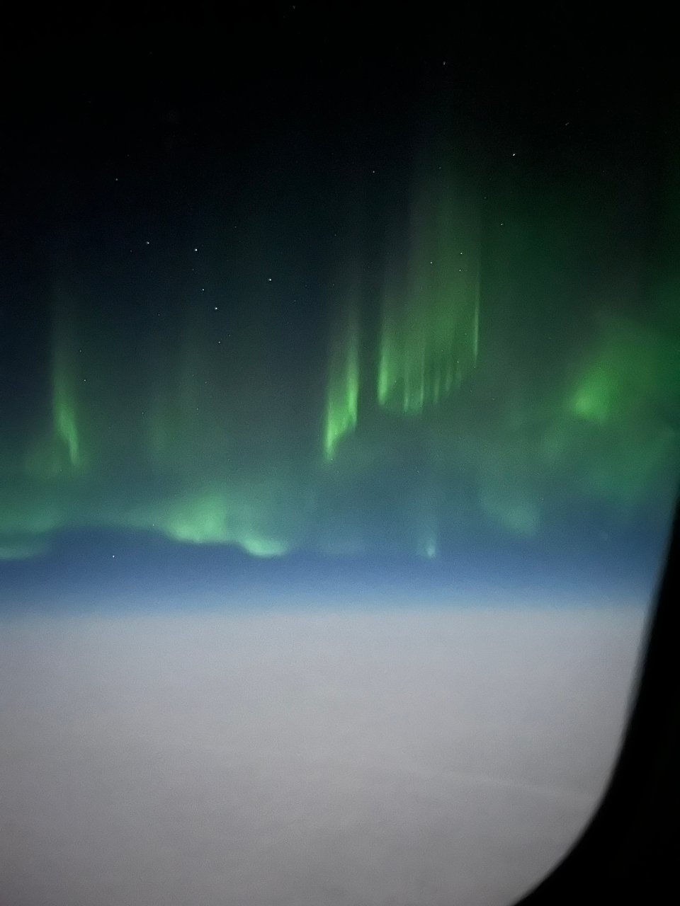 Aurora borealis seen from an airplane window, with green vertical columns and curtains of light above a cloud layer, stars visible in the dark sky above.