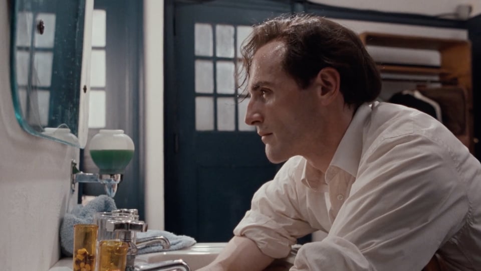Film still from 32 Short Films about Glenn Gould: Colm Feore as Gould leans on a bathroom sink in a white shirt, gazing into a mirror. Pill bottles and a soap dispenser sit by the tap.
