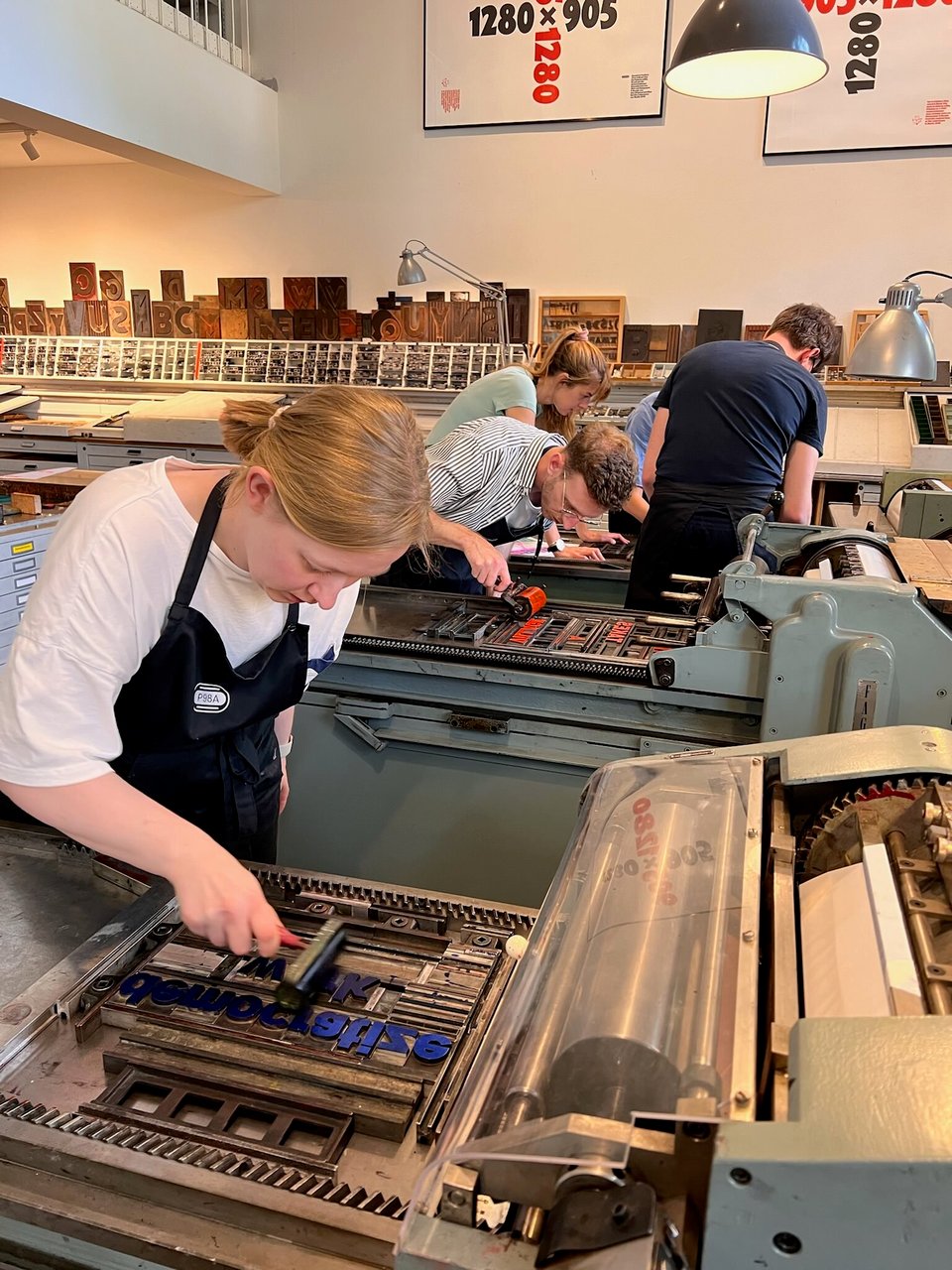 Photo of several letterpress machines and people standing around them, applying paint, for printing poster, everyone appears very focused
