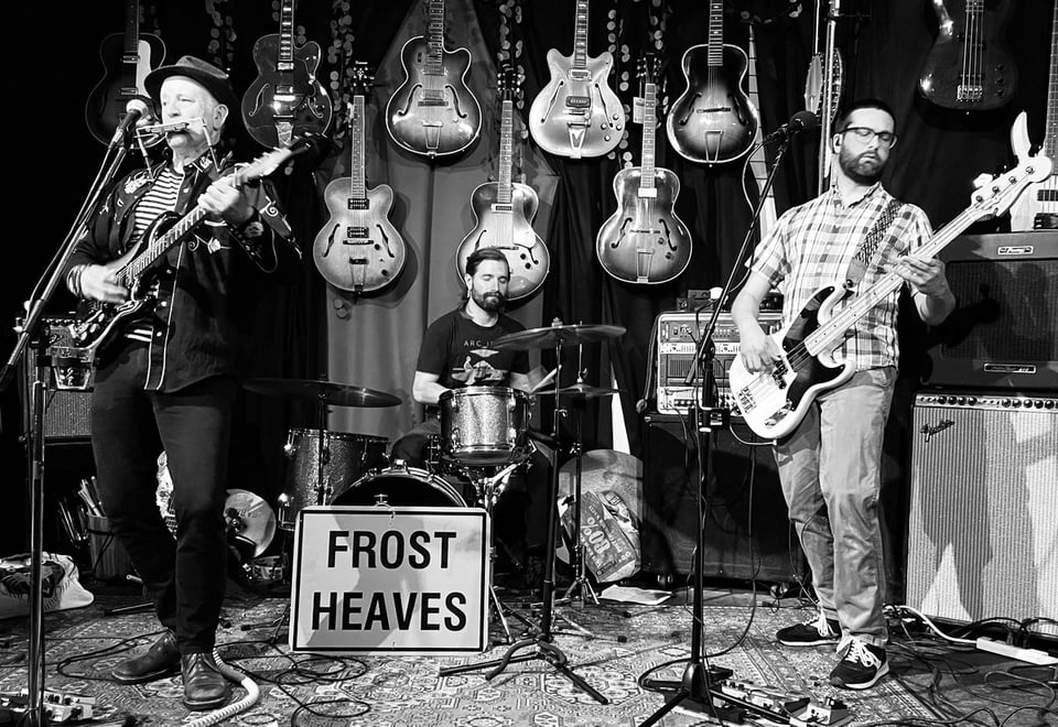A trio of musicians on guitar (with harmonica), drums and bass stand in front of a wall of guitars.