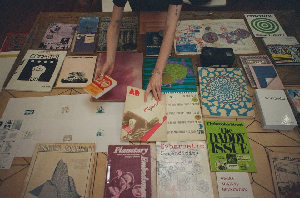 [A close-up of the table with a display of books from the library. A book called Cybernetic Serendipity can be seen, as well as a publication called Radical Software. There's also a calendar, a few publications, Ted Nelson’s Computer Lib/Dream Machines and a Wikipedia book, among others.]