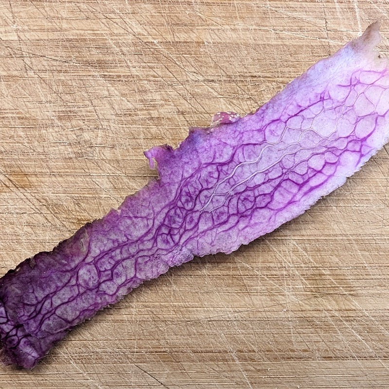 The inside part of a slice of vegetable skin on a scratched bamboo chopping board. The color of the skin goes from black to purple then white. It contains purple veins producing an interesting texture.