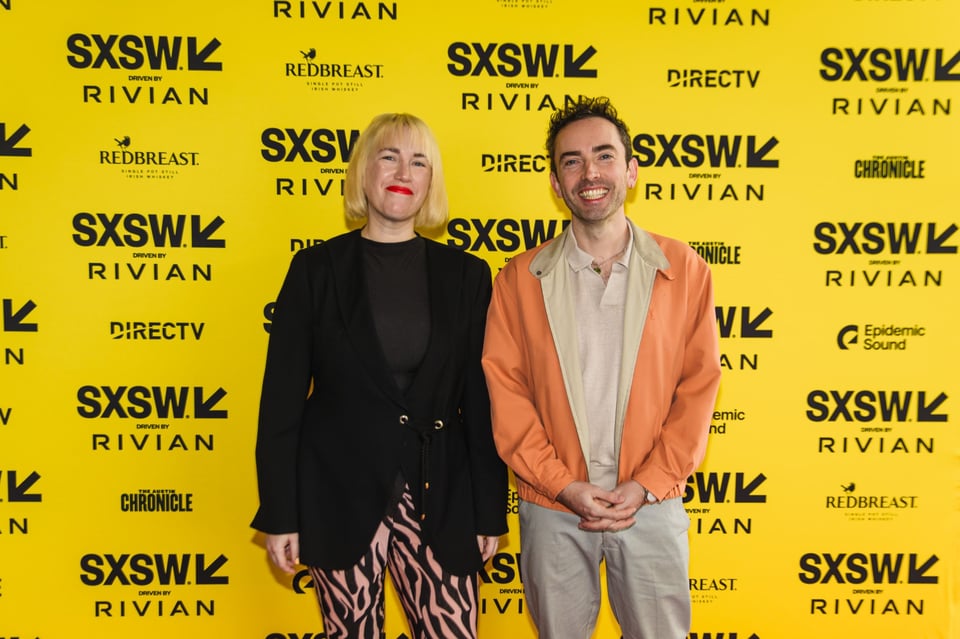 Two people pose in front of a bright yellow SXSW (South by Southwest) step-and-repeat backdrop featuring sponsor logos such as Rivian, Redbreast, and DIRECTV. The person on the left has shoulder-length blonde hair, wears a black outfit with a long blazer and patterned pants, and smiles confidently. The person on the right has short dark hair, wears a light-colored shirt with a peach jacket and grey trousers, and smiles warmly with hands clasped in front.
