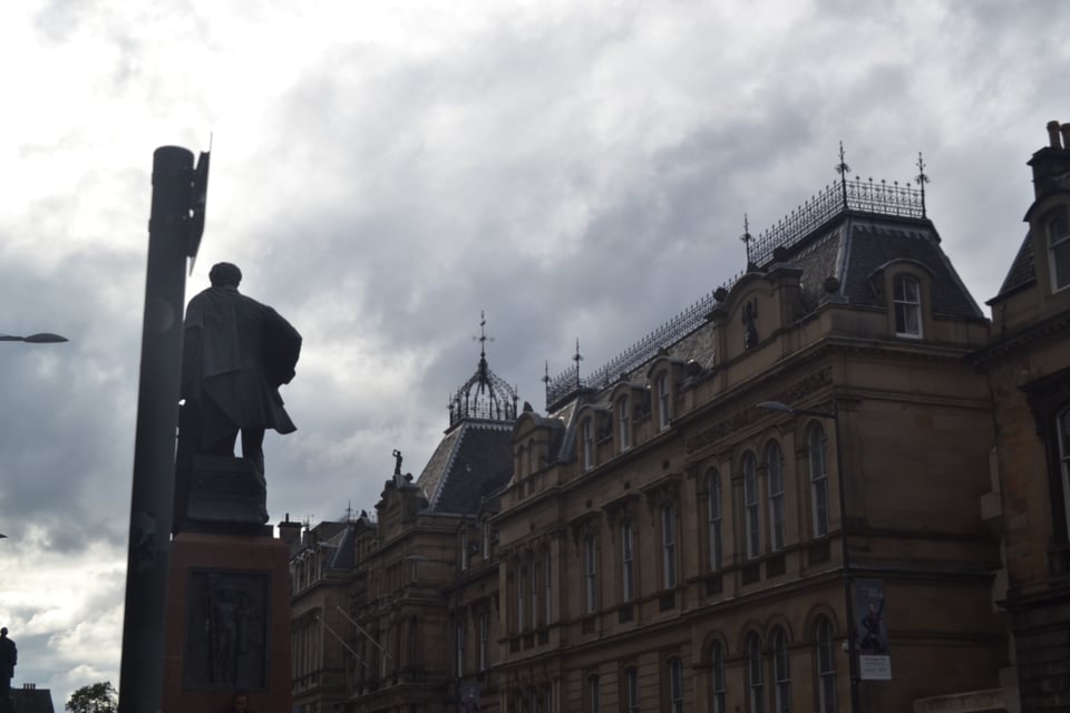 A statue of a man in a coat, his back to the camera. Beyond is a large stone building with lots of windows and a central lantern.