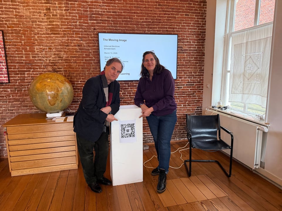 Two people stand next to a podium and point to a QR code.