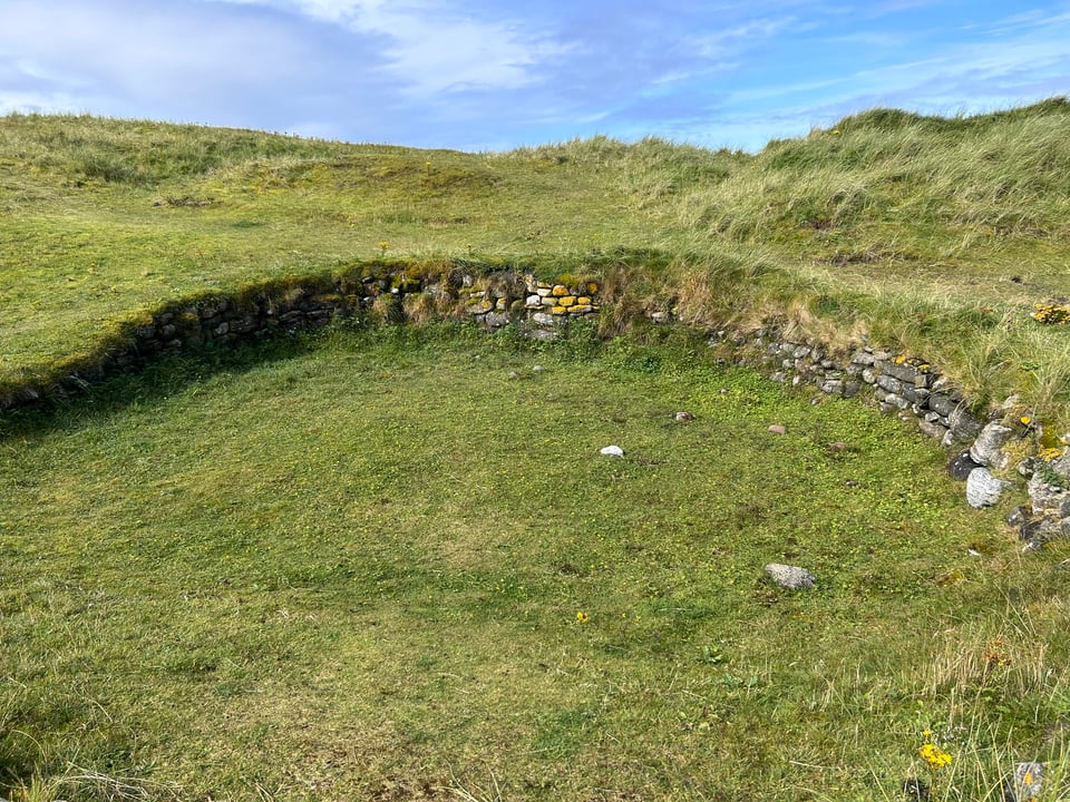 A close up of the drystone wall of one of the Cladh Hallan roundhouses. The walls is embedded in the grassy, sandy ground, turf coming up around it but the stones are clearly visible.