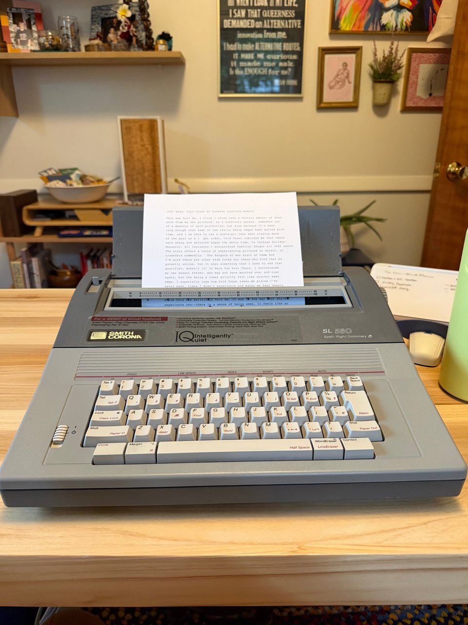 Picture of a gray Smith Corona SL 580 electric typewriter, holding a first draft of JUST READ: COLD TOAST by Kathryn Aldridge-Morris. A white wall with many pictures and a shelf full of books and odds and ends is in the background.