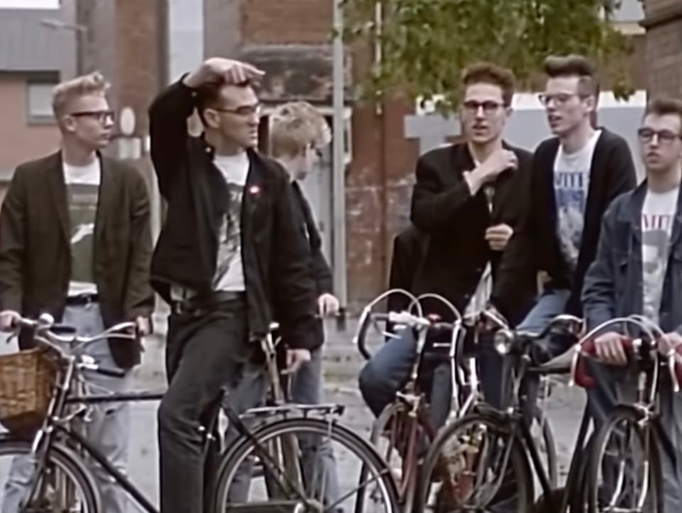 Still from the Smiths video for Stop Me If You Think You've Heard This One Before, showing Morrissey sitting on a bicycle with 5 other people dressed like him sitting on their bikes