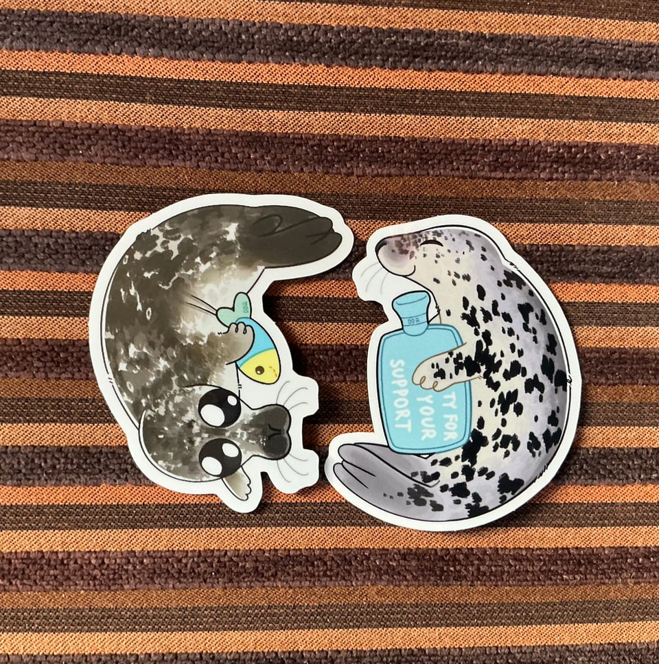 This image shows to stickers with seals drawn in an slightly cartoony way. One is a harbor seal pup with very googly eyes holding a plush fish, the other a lighter colored grey seal pup with closed eyes, holding a blue hot water bottle with the text "ty for your support".