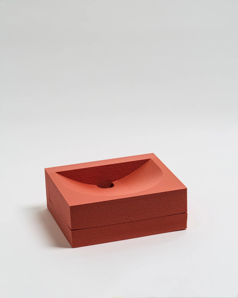 A minimalist red-orange object sits on a plain white surface. It is made from two stacked rectangular layers -- but the top layer features a smooth, concave bowl-like depression with a small circular hole at its lowest point. It looks like abstract art