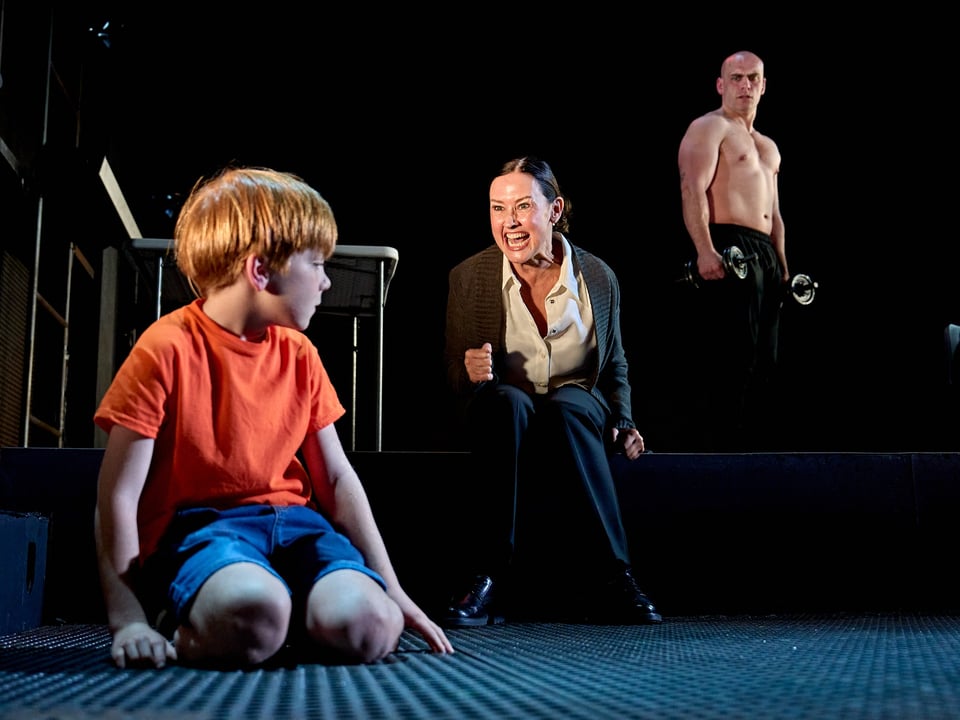 Nathan Jago and Samuel Edward-Cook as Raoul Moat, and Angela Lonsdale as Moat’s mother, in Manhunt. Jago is a young child, kneeled on a grate; he's ginger, wearing a red t-shirt and blue shorts. He's looking away from Lonsdale, who's seated behind him and yelling at him; Lonsdale has her black hair pulled back tight and is wearing a grey cardigan and loose white blouse. Edward-Cook is standing behind both of them, observing the scene. He is shirtless, wearing black trackpants, holding two weights.