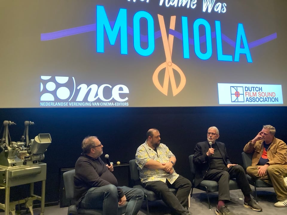 Four men sit in chairs in front of a movie screen with the title "Her name was moviola" projected on the screen. An actual moviola sits in front of the stage to the left of all the men.