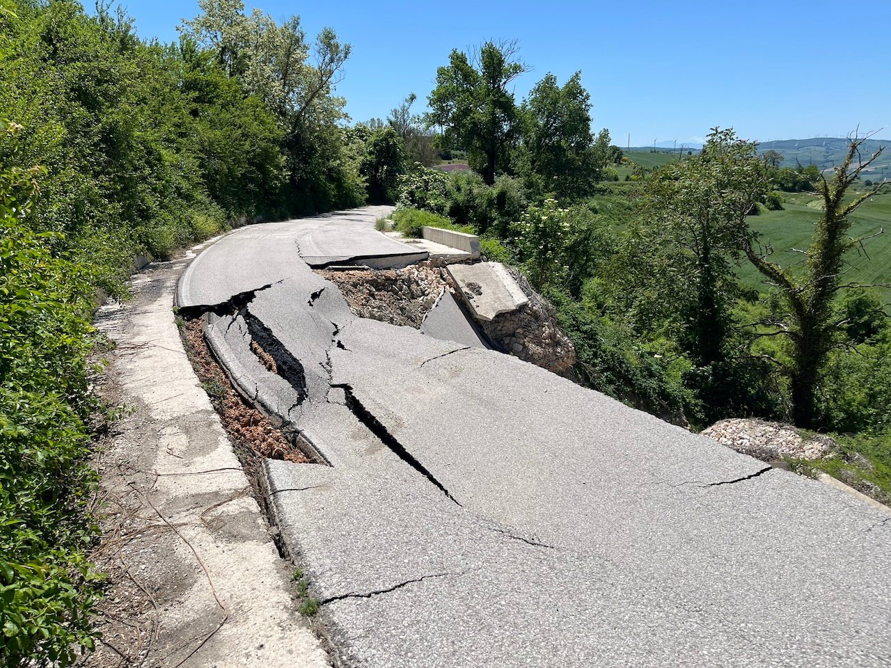 A collapsed country road, its asphalt cracked and sliding down towards the valley. Green bushes line the very narrow and gravelly intact left edge. In the distance, rolling hills with wind turbines under a bright blue sky.