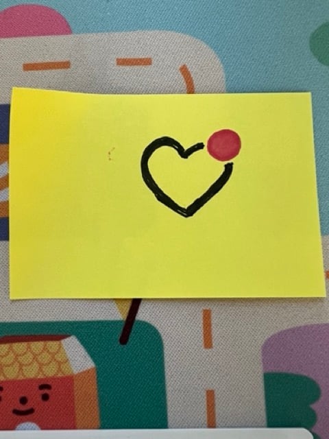 A post it with a black lined heart drawn, and a pink dot on the corner of it.