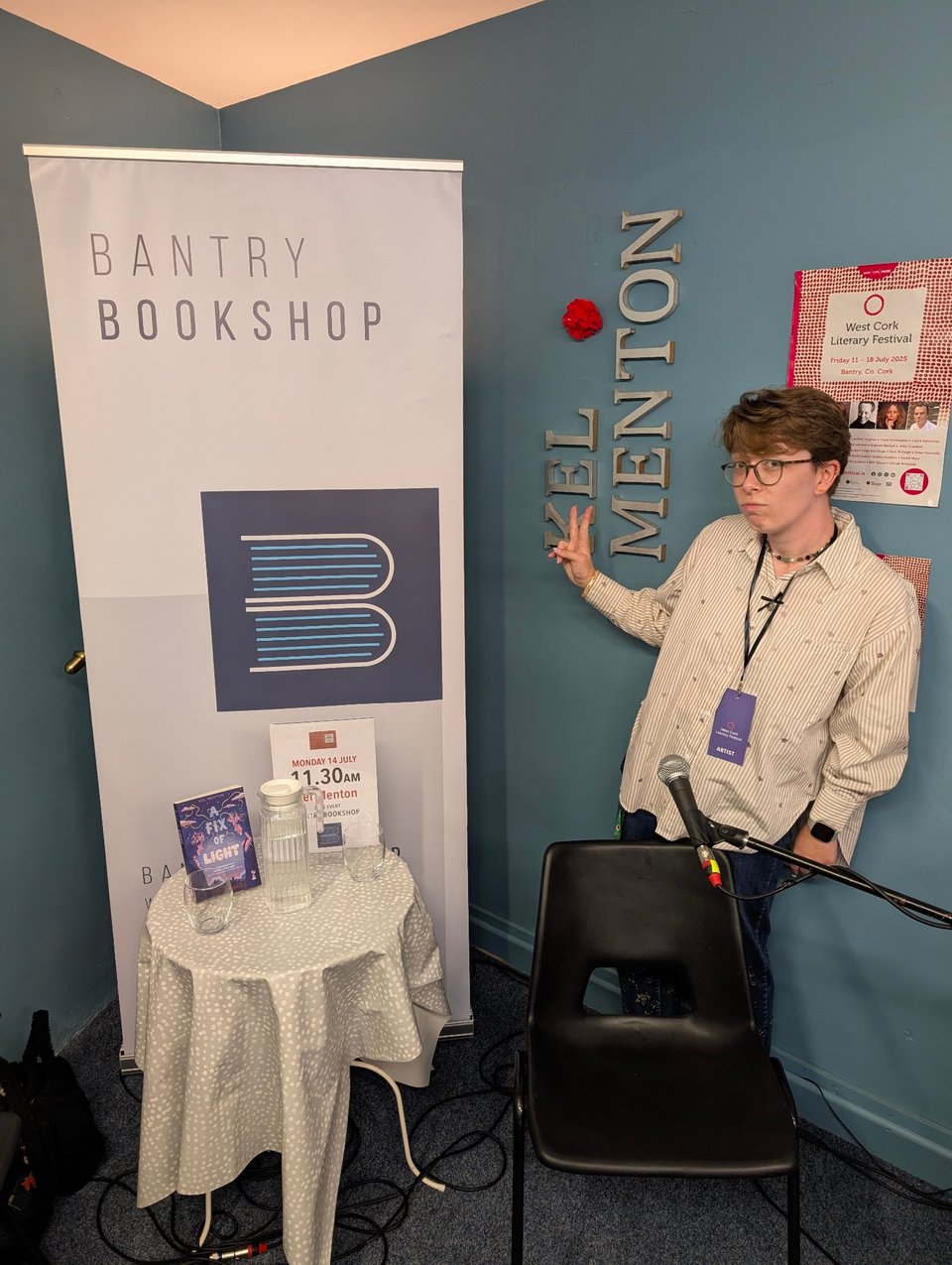 A photograph of Kel Menton. On the left is a banner that reads "Bantry Bookshop". A small table is covered by a paisley tablecloth. On it is a copy of A Fix of Light, a glass jug of water, and two glasses. Kel stands behind a black plastic chair. They hold up a peace sign beside where their name is displayed on the wall. Their lips are pursed.