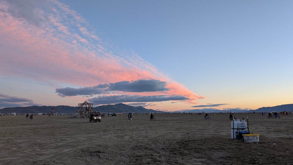 A pink and blue sunset with soft grey clouds over an open, recently-wet desert landscape. Art is visible in the background.