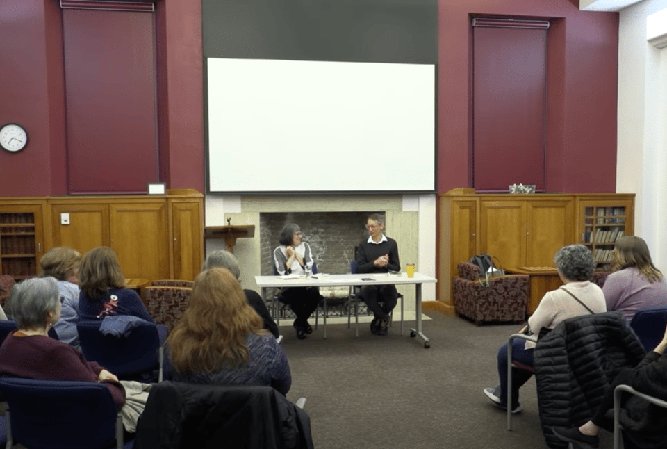 Shot of TV recording of my book event with Susan Ito. We are seated next to each other at a table in front of a large hearth. Audience members are visible in the foreground.