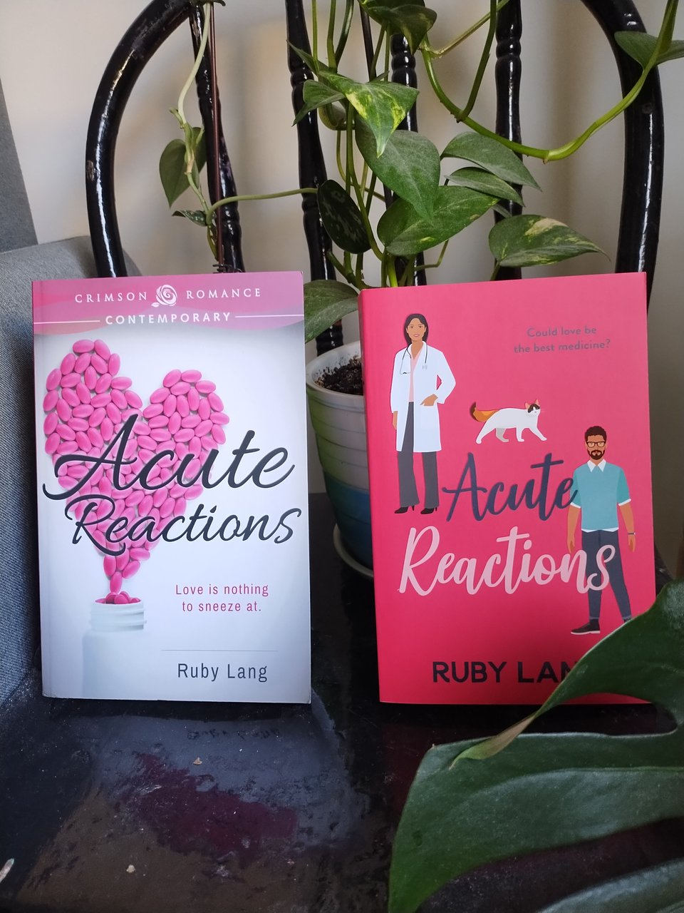 Two copies of Acute Reactions in front of a plant pot: 1. old Crimson cover: white background, pinks pills in heart shape. Cursive title. tag: Love is nothing to sneeze at. 2. Canelo illustrated cover: dark pink background, South Asian woman with dark hair, white coat and dark-haired man with glasses and beard, plus a fluffy cat, cursive title, tagline: Could love be the best medicine?