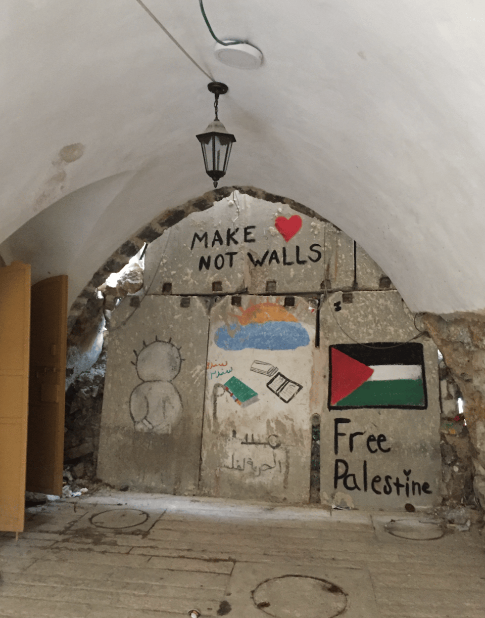 A painted stone wall inside a stone room, including a drawing of a well-known Palestinian cartoon figure (Handala), the words Make {heart} Not Walls, and a Palestinian flag with the words Free Palestine
