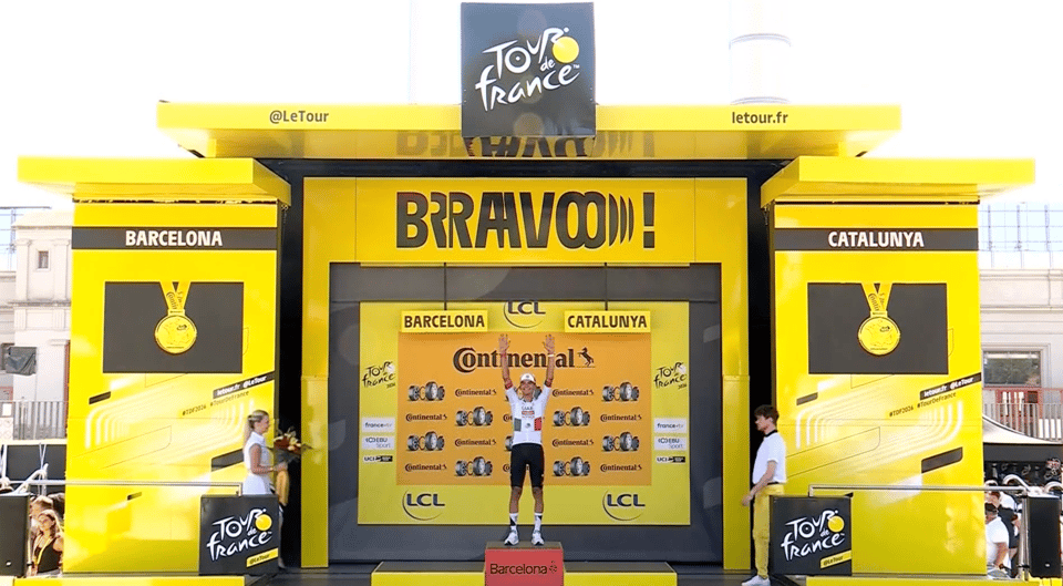 A cyclist stands triumphantly on a podium adorned with yellow decorations, celebrating their achievement at the Tour de France event in Barcelona, Catalunya.