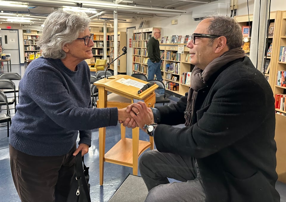 Photo of a white woman and a white man shaking hands in a bookstore, with a look of genuine affection on their faces