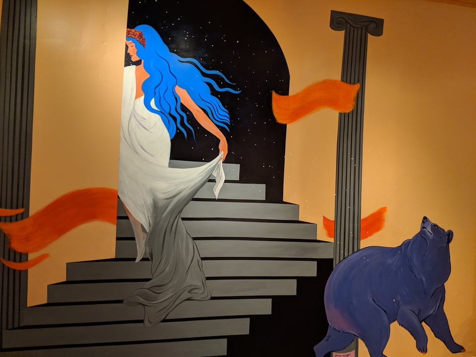 A woman climbs a gray staircase flanked by orange walls while a blue bear watches