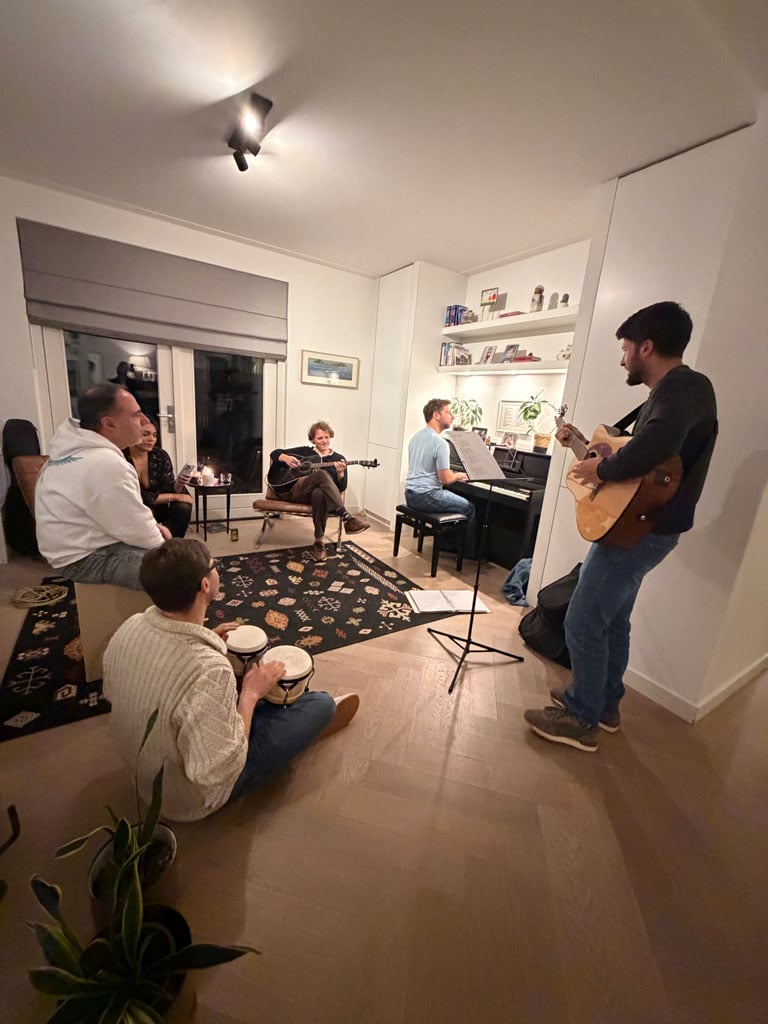 Six people sit and stand around a living room area and make music together - playing the bongos, piano, guitar and singing.