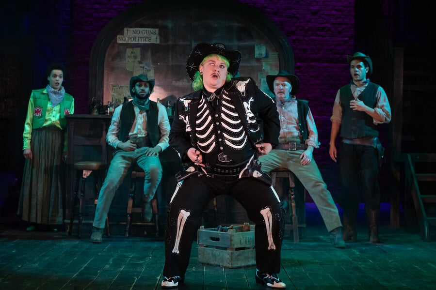 In the centre of the frame is LJ Parkinson, green mullet, pencil moustache, one blind eye, a black leather outfit with white ribs and leg bones and skull decals. Behind LJ, four performers - Lee Braithwaite in a bright green vest and cowboy shirt and dull green skirt, and Michael Elcock, Colm Gormley and Julian Moore-Cook in matching dirty cowboy gear, watch in awe.
