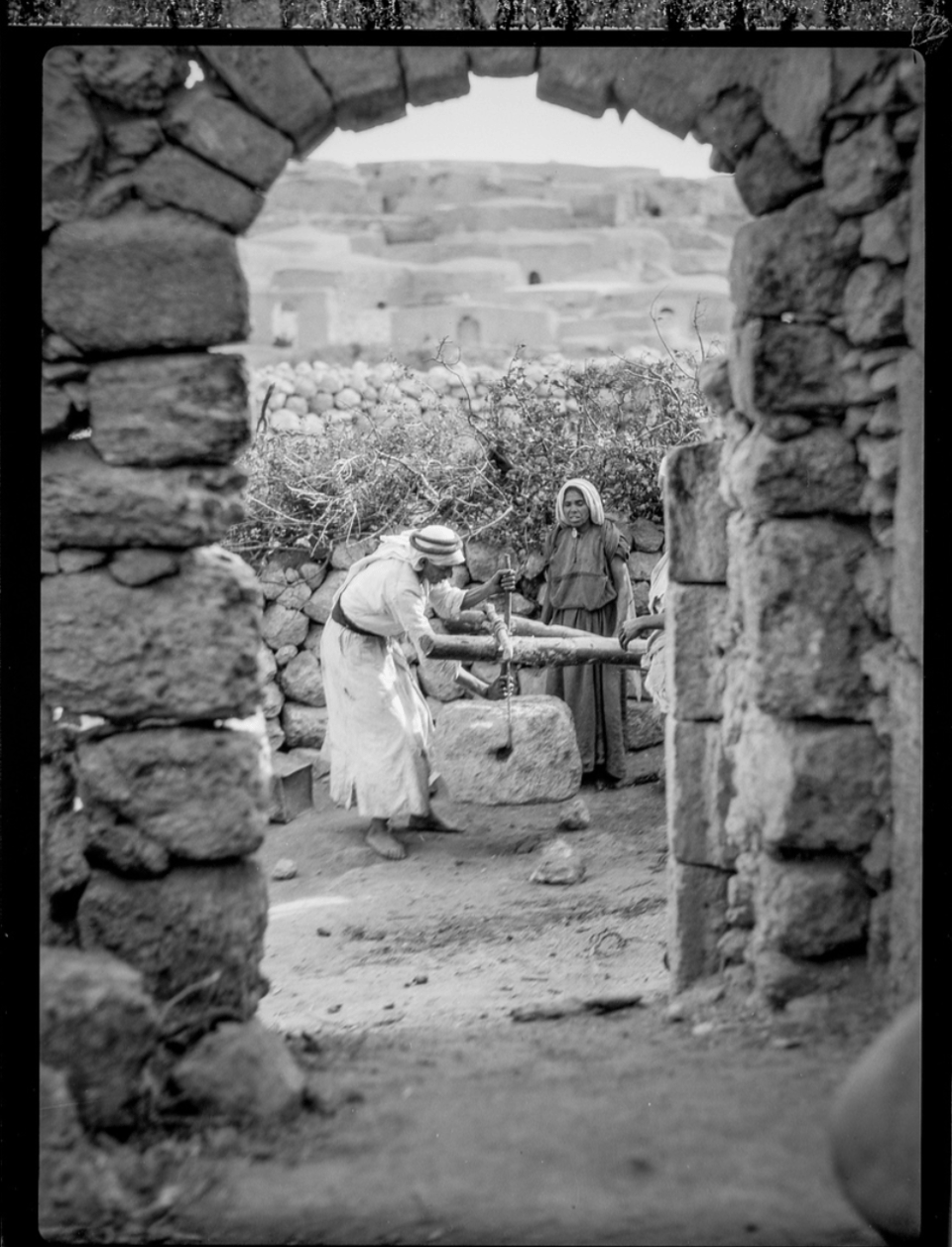 Behind a wooden arch, two people in traditional dress working with a stone press