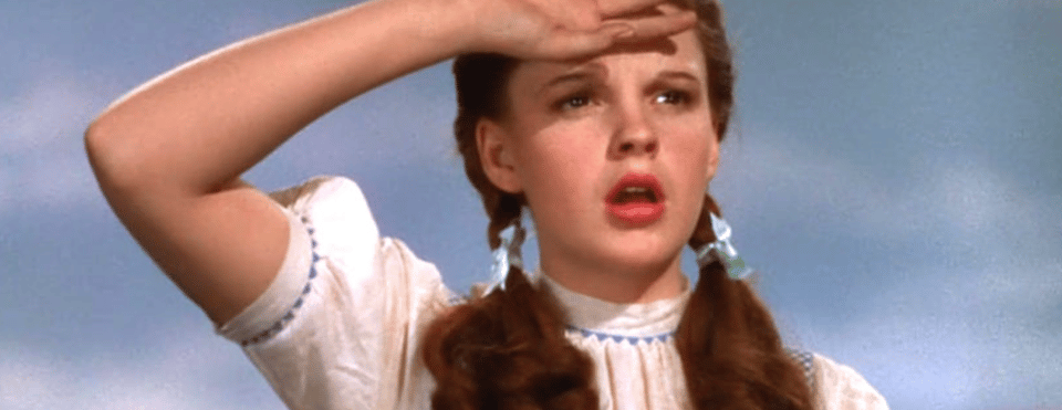 A screenshot from The Wizard of Oz. Judy Garland as Dorothy shades her eyes as she looks into the distance.