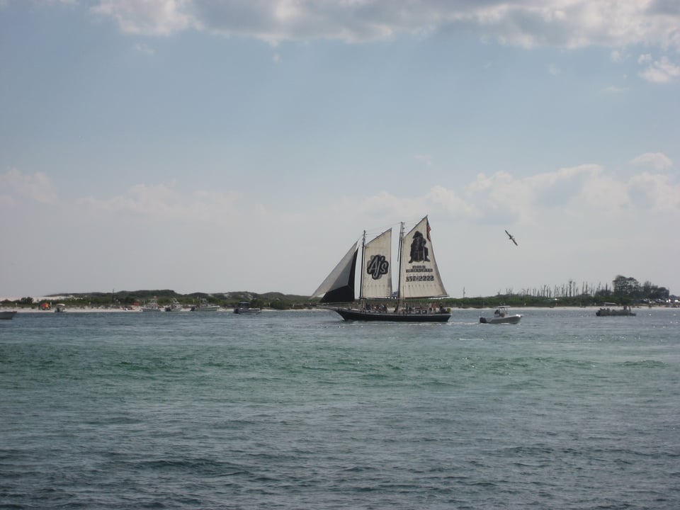 A schooner rigged sailing ship at anchor in shallow water, with a beach beyond. The front sail is black and the others have black graphics on them.