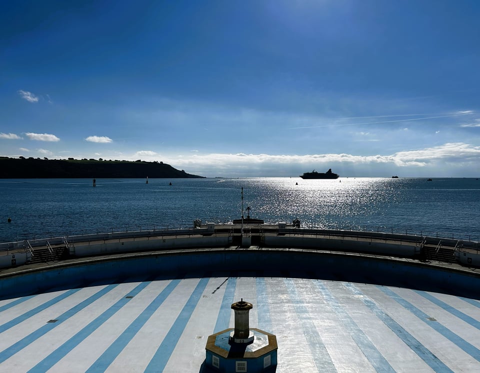 Looking over Tinside Lido to Plymouth Sound. The bold blue sky is making the sea blue, sparkling blindingly bright. In the distance a large cruise ship is silhouetted against the horizon, and a headland edges in on one side. In the foreground is an empty semi-circular swimming pool with a white curving seawall. There's a fountain in the middle, and the floor is painted with two tone blue stripes.