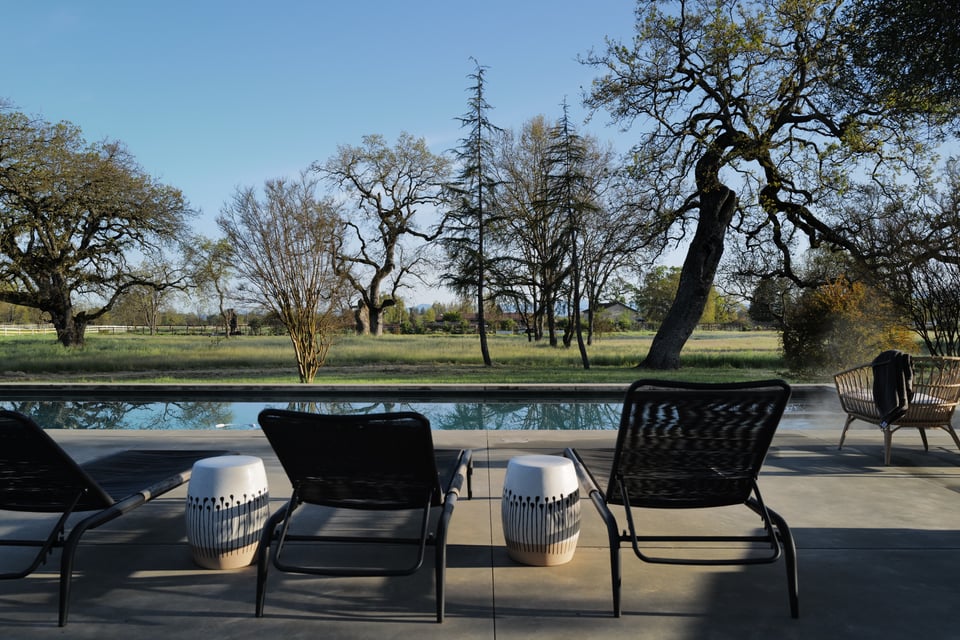 A serene outdoor scene featuring a swimming pool in the foreground, surrounded by a concrete patio with black lounge chairs and decorative ceramic stools. Beyond the pool, a grassy field and leafless trees stretch toward the horizon, bathed in soft late afternoon light under a clear blue sky. The setting suggests a quiet rural retreat, ideal for relaxation.