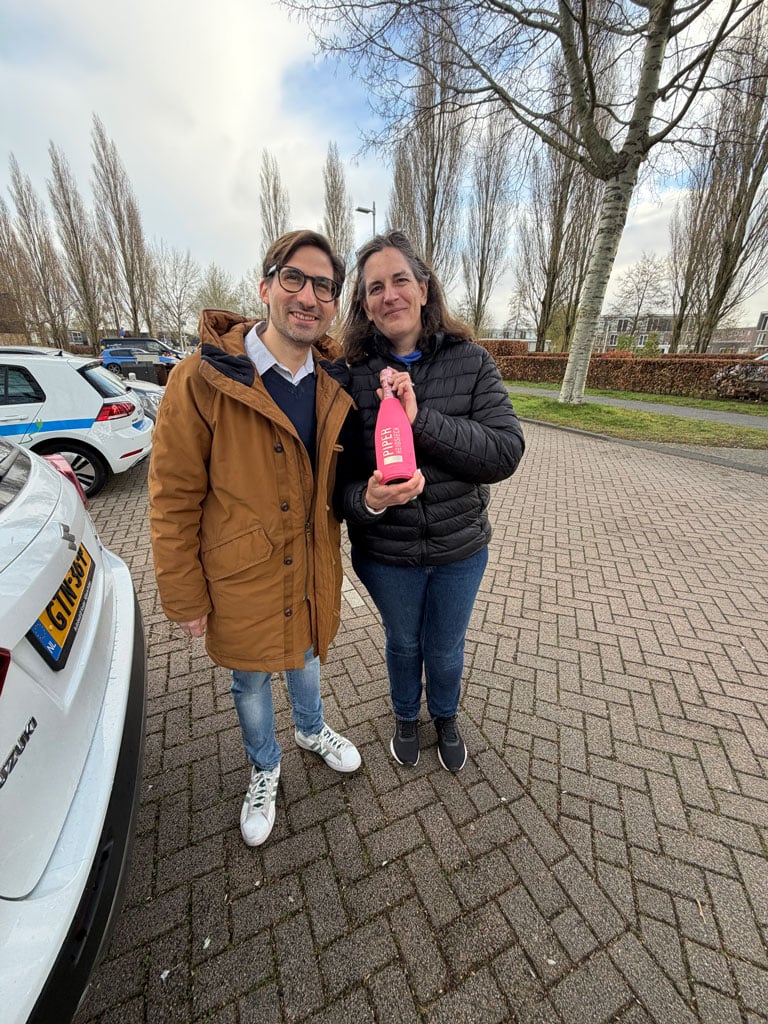A man and a woman stand near a white car smiling and holding a pink bottle of champagne.