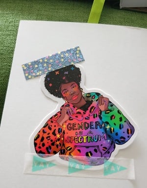 A sticker taped on the inside of a greeting card. The sticker is an illustration of a Black person in a rainbow leopard print hoodie. The front of the hoodie says, “Gender is a spectrum.”