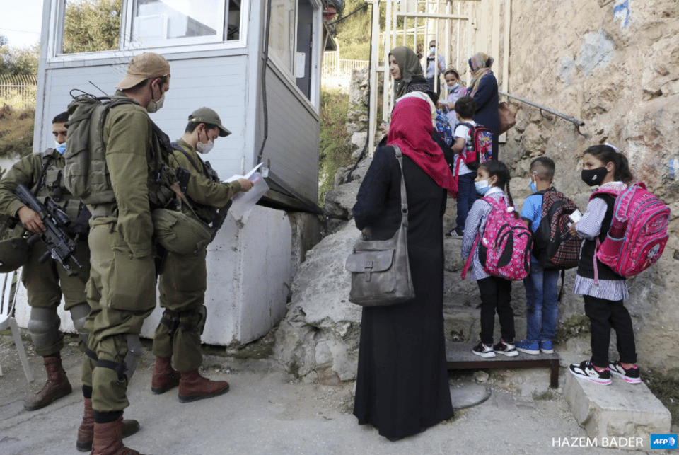 Children with backpacks and a woman with a hijab passing a booth with three soldiers; one is reviewing papers, two have machine guns