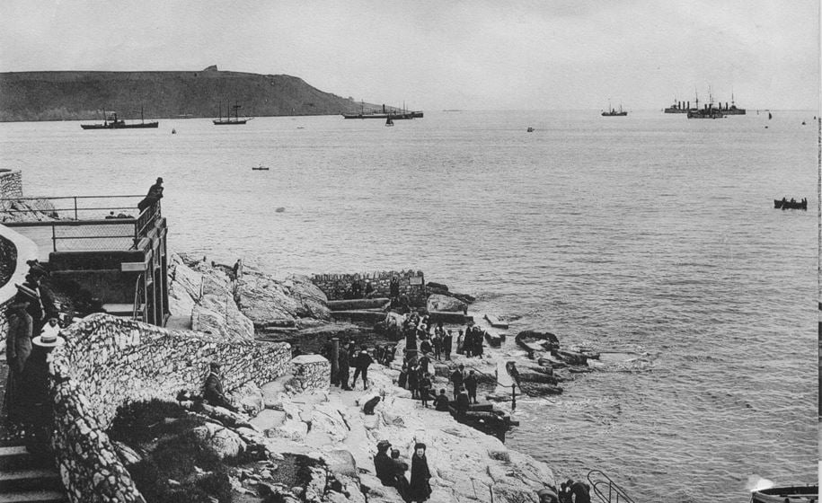 Plymouth Sound with the foreshore in the foreground. People in Edwardian clothing are standing on the rocks and the roof of a small square block.